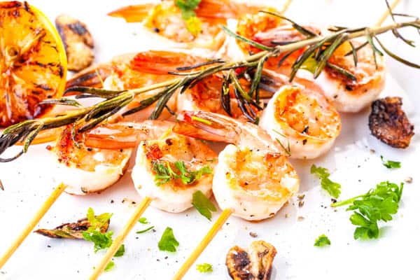 Grilled shrimp on wooden skewers on a plate with rosemary and fresh parsley on top.