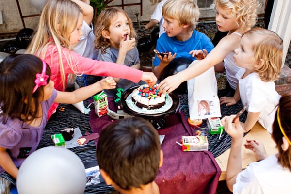 A group of kids around a party table reaching for the cake. 