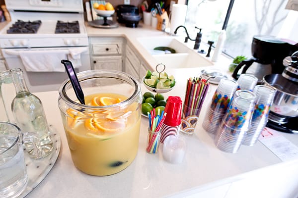 Large jar of sangria on a counter next to cups and straws for a party. 