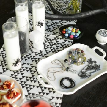 A white tray with jewelry making items for a Halloween DIY party next to white pillar candles with giant bugs on them.