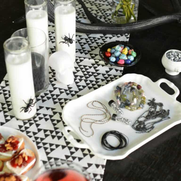 A white tray with jewelry making items for a Halloween DIY party next to white pillar candles with giant bugs on them.
