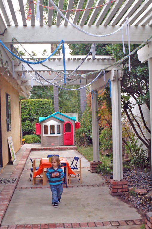 A little kid in a backyard with streamers hanging from the patio. 
