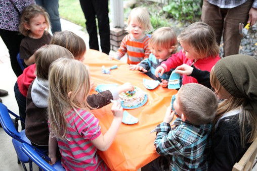 Kids sitting around a table at a party with a cake in the middle. 