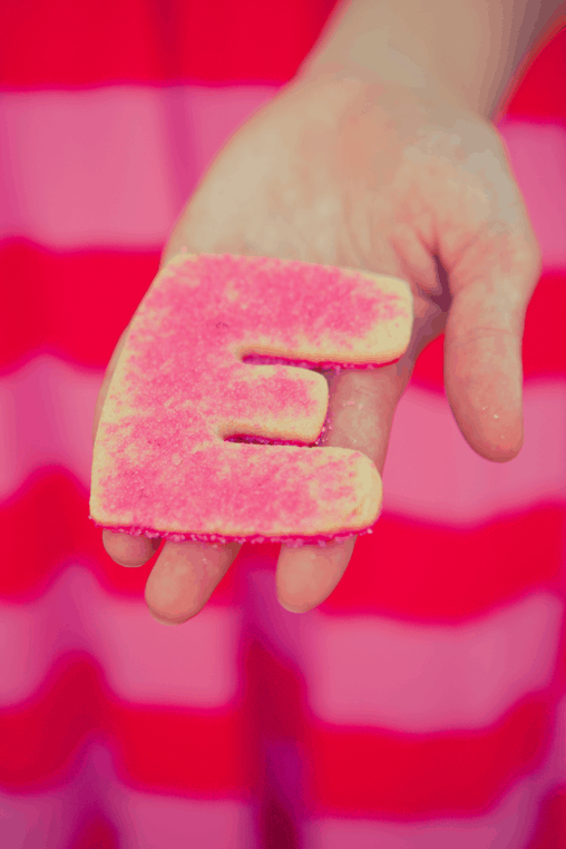 A woman's hand holding an "e" shaped sugar cookie with pink sanding sugar. 