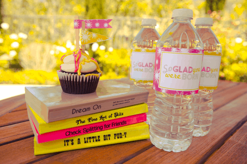 A stack of painted books with a cupcake on top next to bottles of water. 