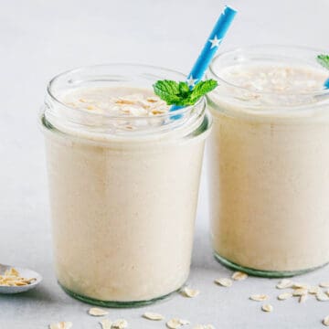 Banana Oatmeal Smoothies in glasses with oats sprinkled around, mint garnish and blue straw with stars on it.