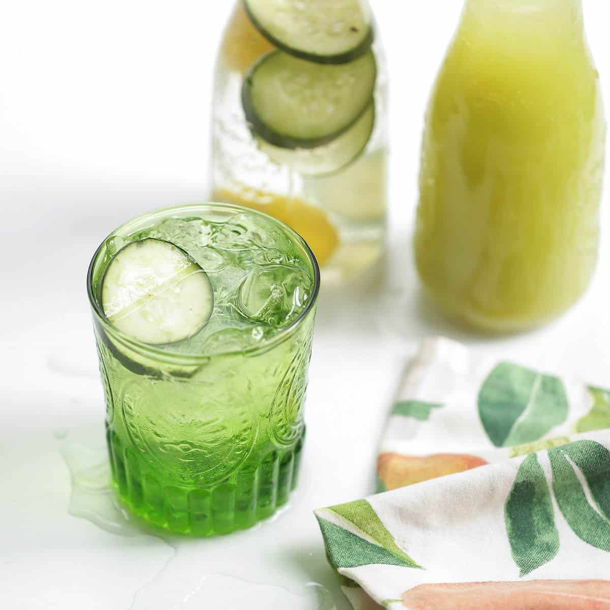 2 pitches on a table, one with green juice and one with water and cucumbers next to a green glass on a table.