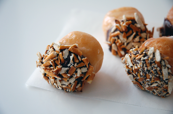 Three donut holes dipped in chocolate and crushed pretzels on a piece of white wax paper on a white table. 