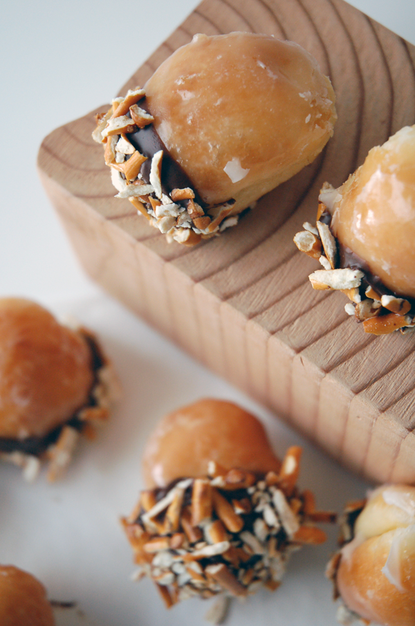 Close up of 2 donut holes that have been dipped in chocolate and crushed pretzels on a wooden block with more donut holes on the table below. 
