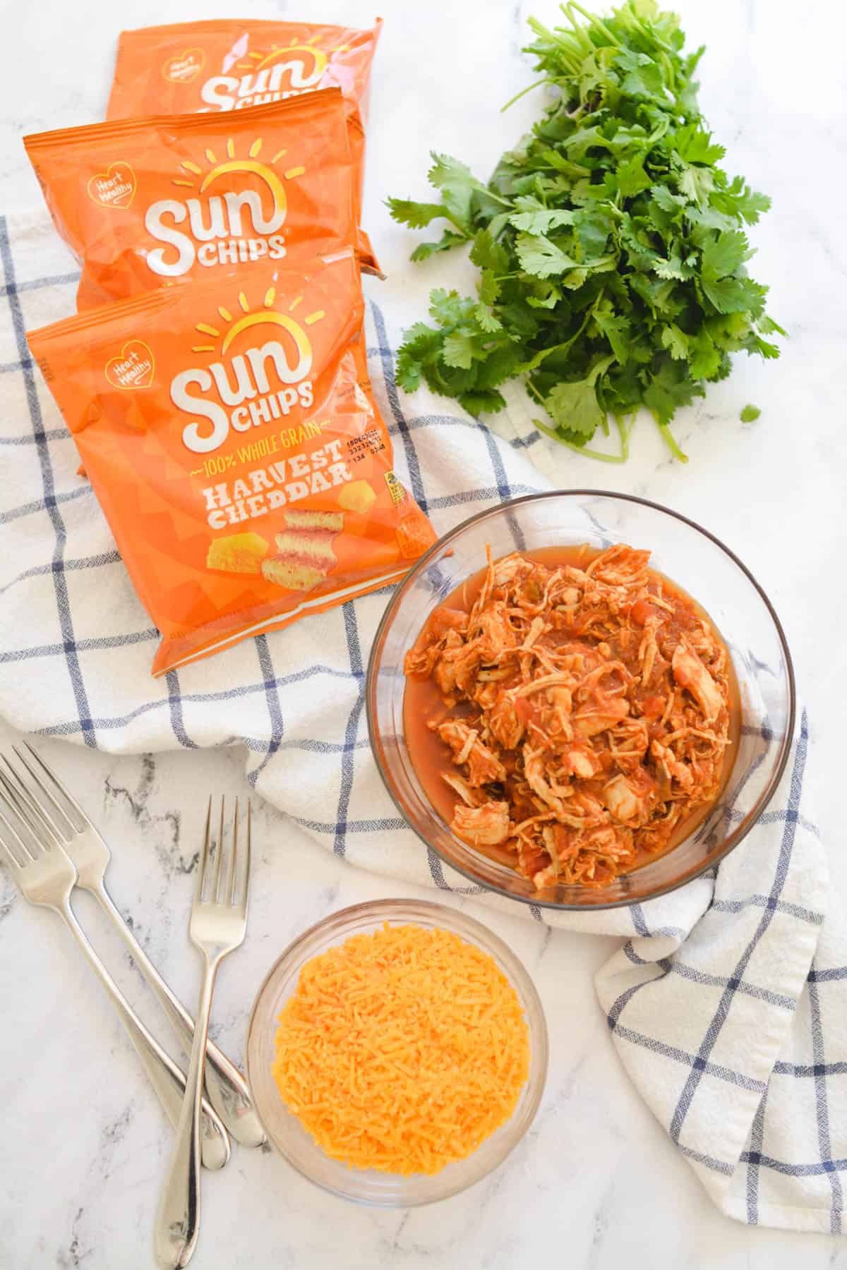 A bowl of salsa chicken on a table next to cilantro, cheese and bags of Sun Chips.