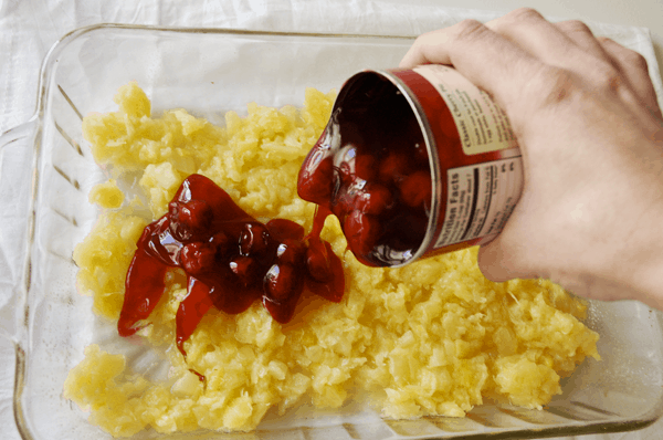 A glass dish with crushed pineapple and a girl pouring a can of cherry pie filling over it to make our favorite easy homemade Cherry Pineapple Dump Cake recipe. 