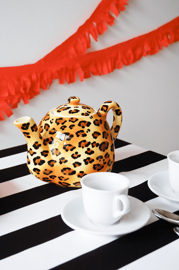 Close up of a leopard teapot on a table covered in black and white striped fabric. 