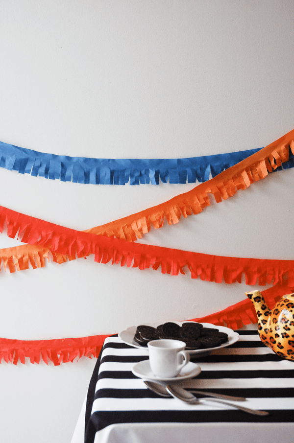 Close up of fringed garland on a wall behind a small table with a plate of cookies. 