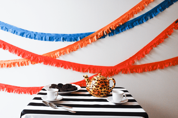 Colorful fabric fringe streamers on a wall behind a small table with a leopard print tea pot and a plate of cookies. 