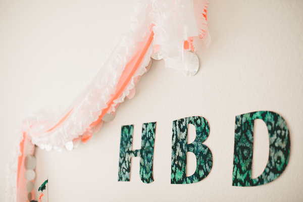 A wall sign that says "HBD" with ruffled streamers from plastic tablecloths on the wall next to it.