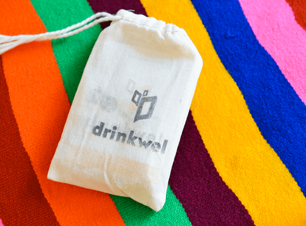 A small muslin bag that says "Drinkwel" on a rainbow striped blanket. 