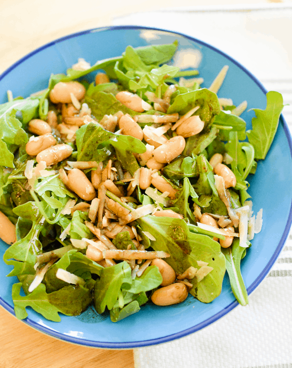 Arugula in a bowl topped with white beans and parmesan cheese. 