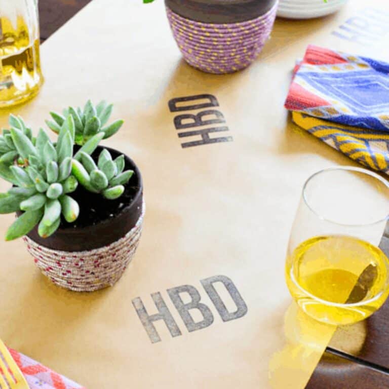 Kraft paper runner on a table stamped with "HBD" next to wine glasses, napkins and potted plants for a party.