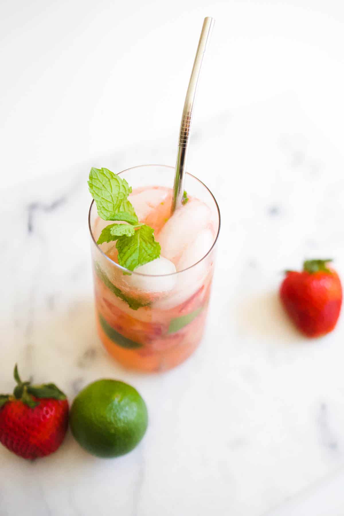 A glass on a table next to a lime and 2 strawberries with infused sparkling water and metal straw.