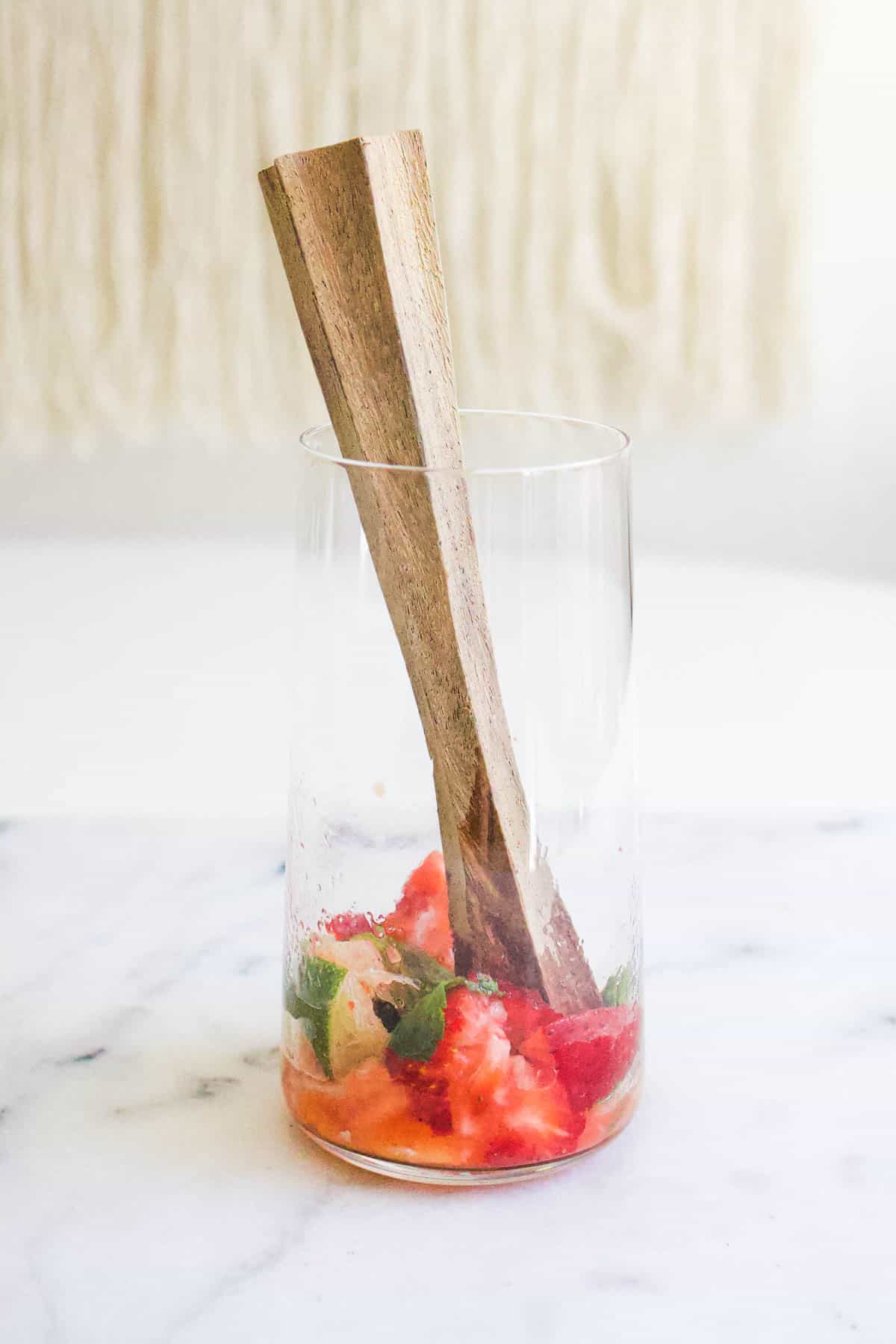A muddler sticking out of a glass with strawberries mint and ginger muddled in the bottom.