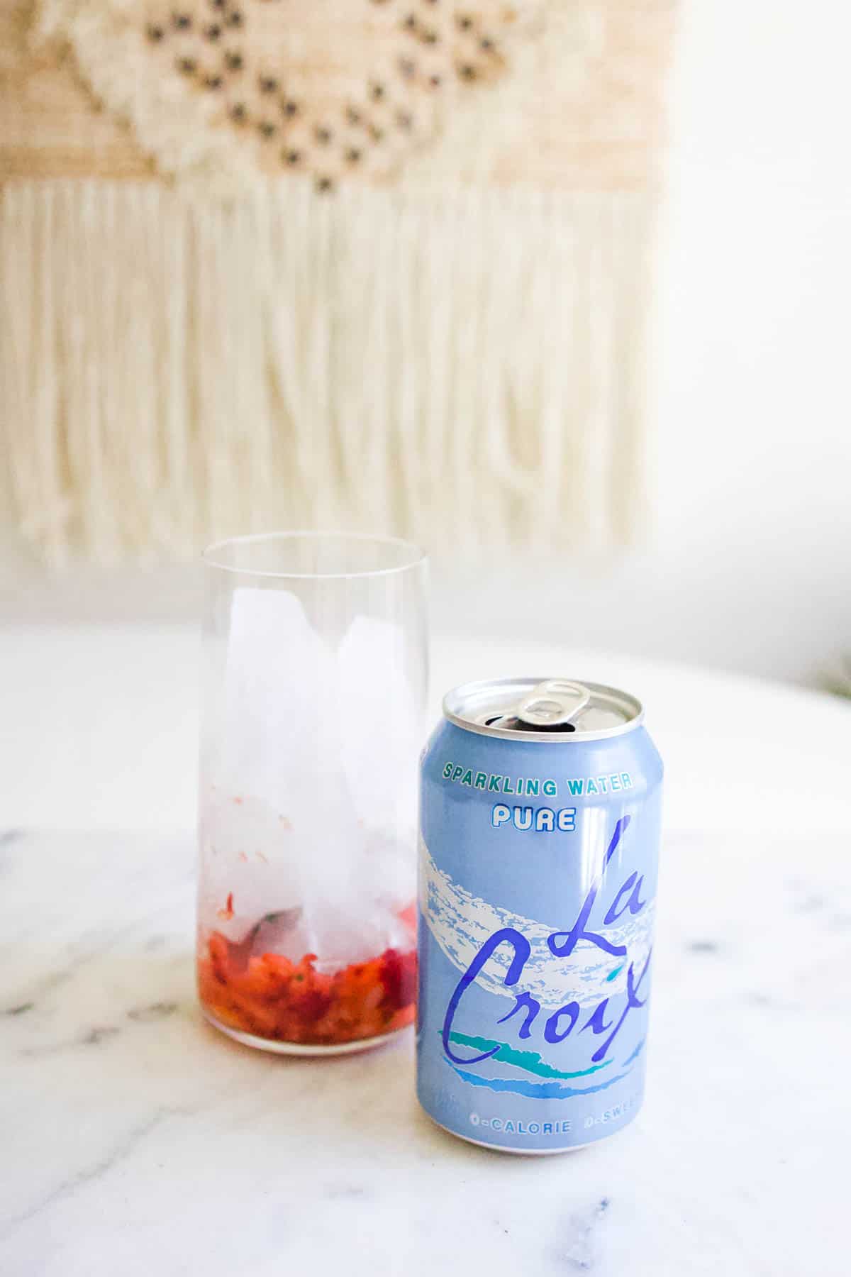 A glass with muddle strawberries next to a can of sparkling water.