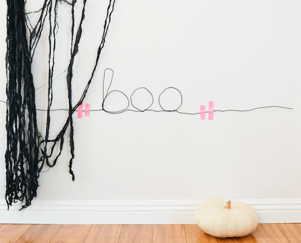 DIY wall sign made out of wire that says "boo" with a white pumpkin on the ground and a spooking black fabric hanging on the wall