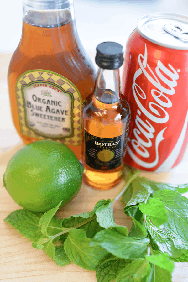 Ingredients including a can of Coke, a mini bottle of rum, a bottle of agave, fresh lime and mint to make a mojito with cola.
