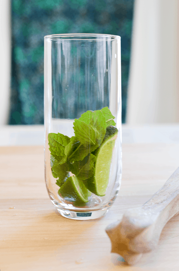 A tall skinny cocktail glass with fresh lime wedges and fresh mint on a counter next to a muddler. 