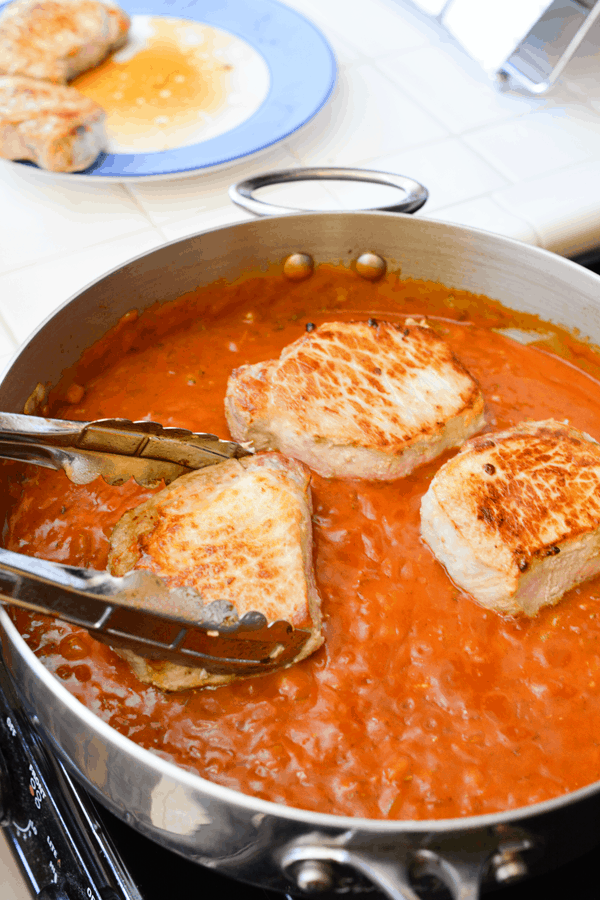 Browned pork chops being added to a simmering pan of red sauce.