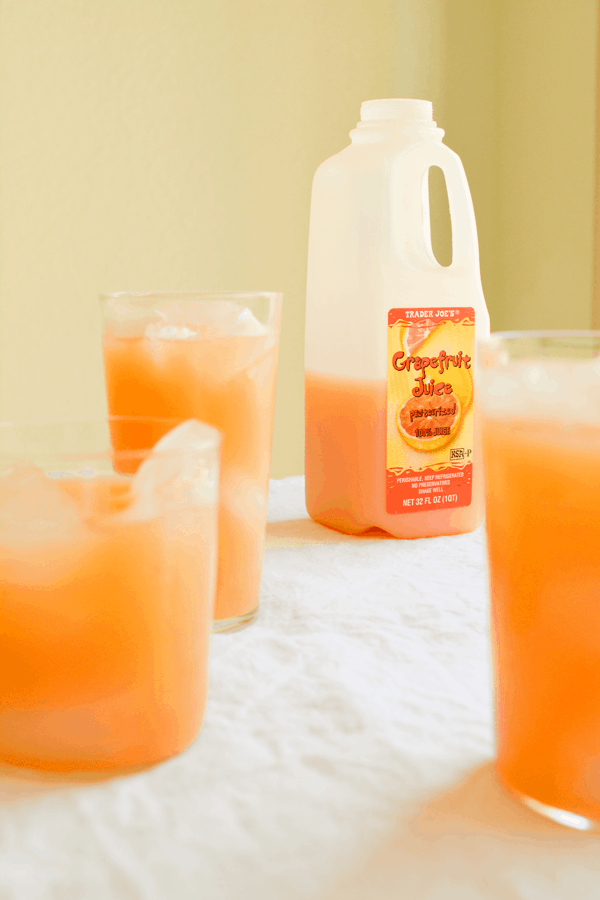 A jug of grapefruit juice from Trader Joe's on a table with white tablecloth with a few glasses of cocktails next to  it. 