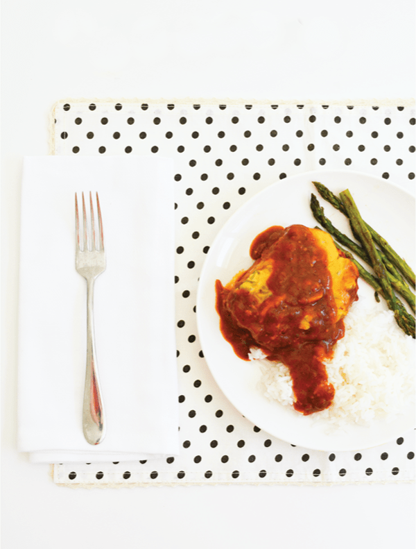 Pork chops in red sauce over rice with asparagus on a white plate on a dotted table mat.
