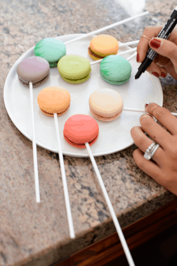 Variety of colorful macarons on a plate with lollipop sticks and girl holding an edible ink pen.