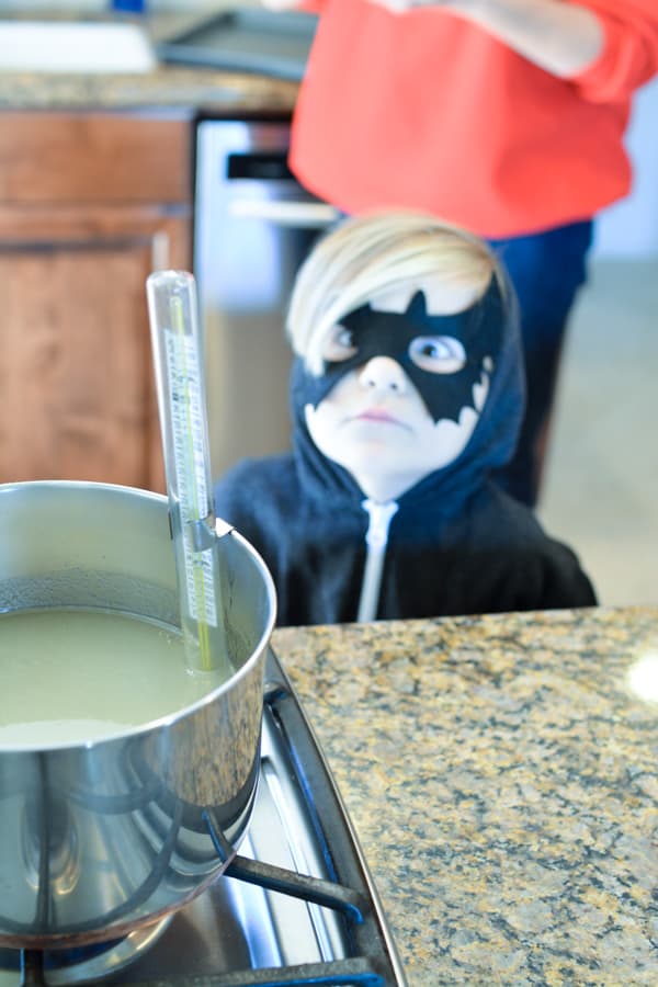 Child in a bat mask standing back from the stove where there is a saucepan of sugar with a candy thermometer in it.