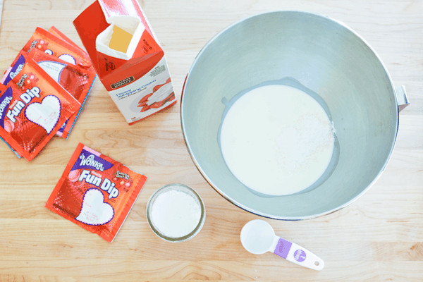 Fun Dip Whipped Cream Ingredients The bowl of a mixer sitting on a wood cutting board filled with heavy cream surrounded by red Fun Dip packets.