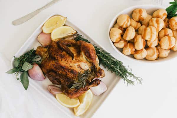 A whole roasted chicken in a baking dish surrounded by fresh herbs and aromatics.