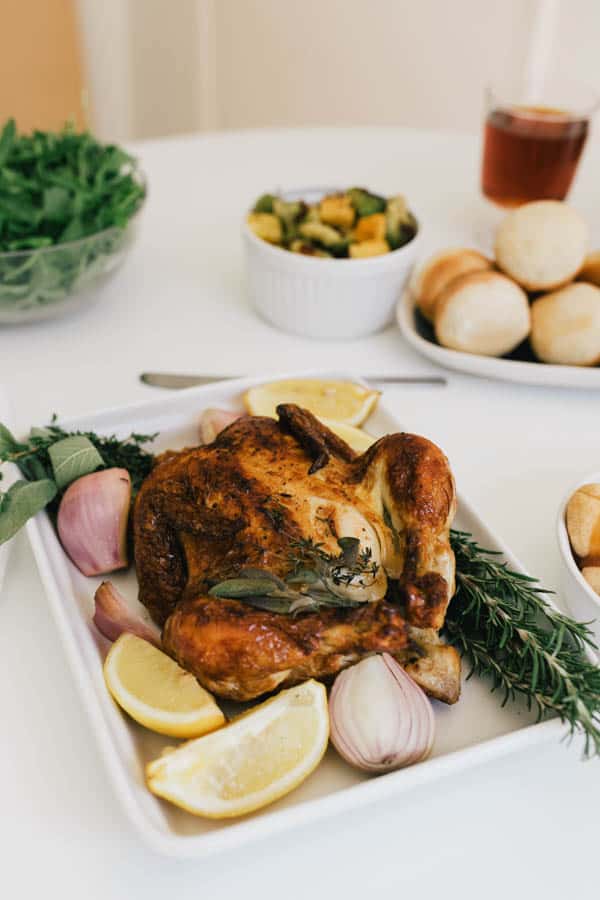 A roasted chicken in a white dish surrounded by lemons, onions and fresh herbs.