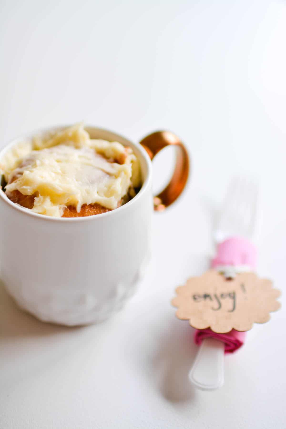 A store-bought cinnamon roll in a coffee mug next to a plastic fork with a gift tag.