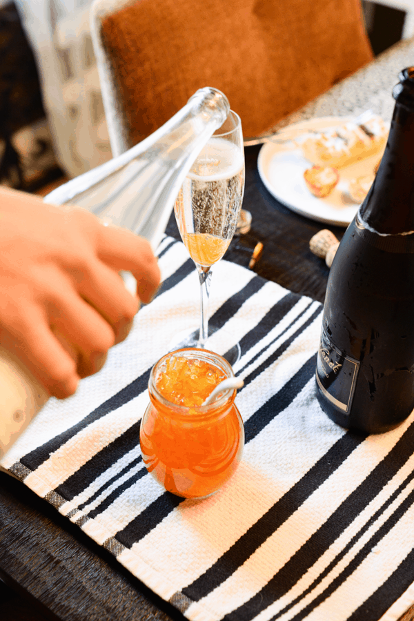 A woman pouring champagne over orange marmalade in a champagne glass with orange marmalade next to a small jar of jam and a bottle of champagne.