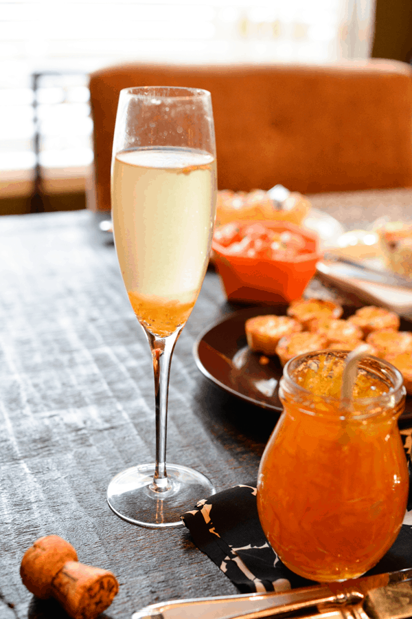 Champagne and marmalade in a champagne flute next to a jar of orange marmalade and several plates of appetizers on a dark wood table.