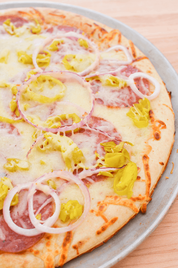 Close up of a baked salami pizza on a pizza pan with melted cheese, red onion slices and pepperoncini. 