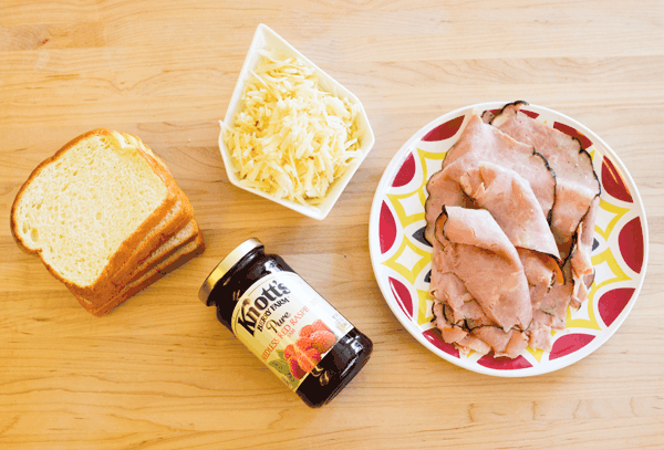 A plate with black forest ham on a wooden board next to a bowl of shredded cheese, a jar of raspberry jam and a stack of King's Hawaiian Bread.