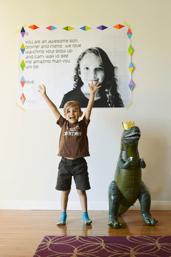 A child with arms raised, next to a dinosaur shaped balloon, in front of an oversized happy birthday banner.
