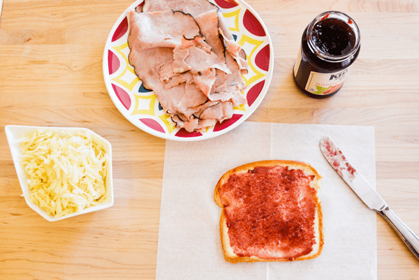 A piece of Hawaiian bread spread with raspberry jam on a piece of white waxed paper on a wooden cutting board next to a knife, a jar of jam, a plate of ham and a bowl of shredded white cheese.