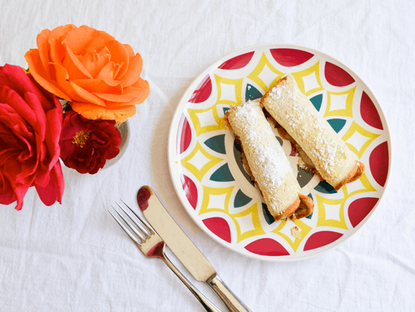 A plate with Monte Cristo roll-ups on a white table next to a vase of flowers and a fork and knife. 