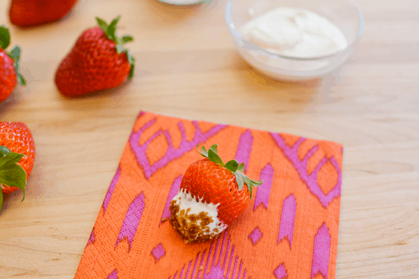 A fresh strawberry dipped in sour cream and then dipped into brown sugar on an orange and pink patterened napkin on a wooden cutting board next to other strawberries.