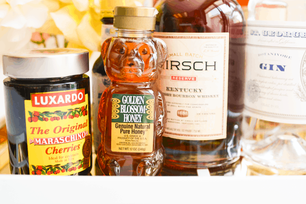 Close up of a bear shaped honey bottle on a bar cart next to a jar of cherries and bottles of liquor.