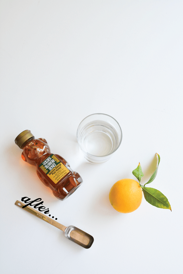 A bottle of honey on a white table next to a measuring spoon a lemon and a glass of water. 