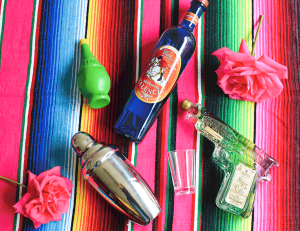 A blue bottle of tequila, a bottle of tequila that looks like a gun, a cocktail shaker, a bottle of lime juice on a striped Mexican blanket. 