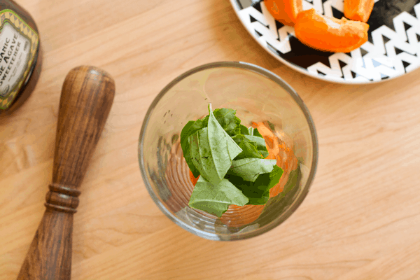 A glass with fresh basil and pieces of tangerine next to a muddler.