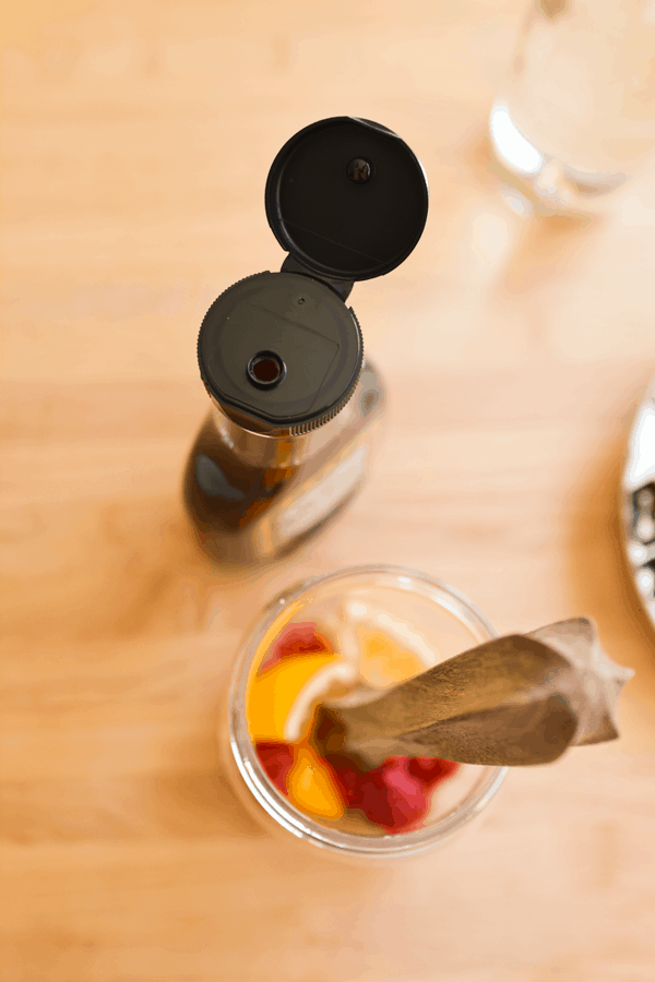 Agave syrup bottle on a wooden board next to lemons and raspberries being muddled together in a glass.